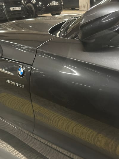 The scene is set in an enclosed multi-story car park in Gillingham, United Kingdom. The primary subject is a dark grey BMW sDrive 320i vehicle, oriented with its front portion facing right from the viewer's perspective. The front left fender and driver's side door of the BMW exhibit multiple superficial linear scratches and scuff marks, appearing light against the dark paintwork, with some concentrated bright white abrasions. The driver's side mirror casing also shows similar scuffs. The vehicle's lower body panels reflect yellow striped markings from the parking surface. In the blurred background, other vehicles are visible, including a dark SUV with the UK registration plate "GM17 AYB" and another dark vehicle displaying "BV22 AZN". Concrete pillars and ceiling structures typical of a parking garage are present. The environment is illuminated by artificial overhead lighting, creating reflections on the car's surface. No individuals or active interactions are depicted.
