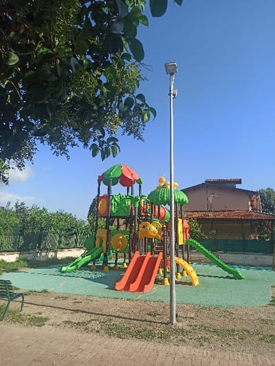 An unoccupied children's playground is depicted under a clear blue sky in San Marzano sul Sarno, Italy. The central feature is a multi-level, colorful plastic and metal play structure, predominantly green, red, yellow, and orange. It includes two parallel red slides, a curved green slide on the left, and a straight green slide on the right, along with various platforms, climbing elements, and mushroom-shaped canopies. The ground directly beneath the play structure is covered with a light green, possibly rubberized, safety surface.
In the foreground, a dirt path with sparse grass traverses the bottom of the frame. A green metal park bench is partially visible on the far left. A tall, slender silver light pole, equipped with a rectangular LED light fixture and an auxiliary sensor box, stands prominently in the mid-right.
The upper left quadrant is occupied by the overhanging branches of a tree with dark green leaves and numerous small green berries or buds. In the background, behind the playground, a green chain-link fence borders an area dense with green foliage and trees. To the far right, a two-story building with a reddish-brown tiled roof and light-colored walls is partially visible. The scene is illuminated by direct sunlight, casting shadows. No individuals are present.