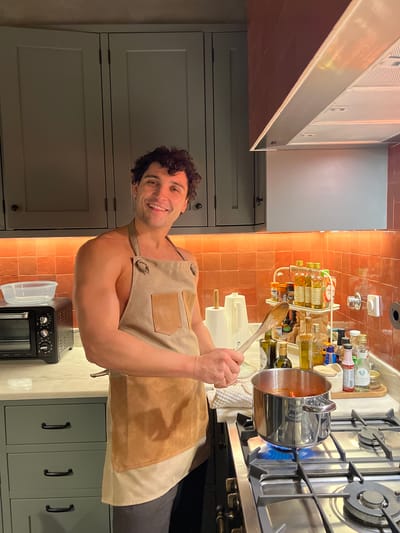 A man in a tan apron is cooking in a kitchen with orange tile backsplash. He is stirring a pot of red sauce on a gas stove, looking towards the camera and smiling. The kitchen includes overhead cabinets, a stainless steel vent hood, a countertop with bottles and containers, a microwave oven, and lower cabinets. The location is Gondarém, Portugal.