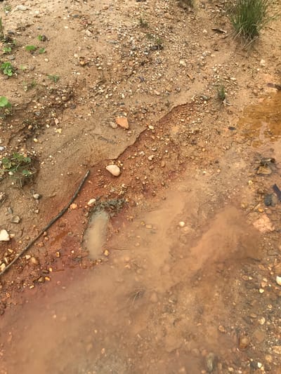 The image shows a section of muddy ground in an indeterminate outdoor location. The ground consists of sand, gravel, small rocks, and soil with some sparse vegetation. A pool of muddy water is present on the right side of the image. A small stick is partially submerged in the mud. No people or animals are visible. The specific location within the city of Ietarek Dua, Indonesia cannot be determined from the image.