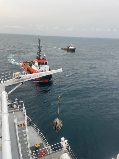 An elevated view from a vessel's deck captures a maritime scene off the coast of Orimedu, Nigeria. In the immediate foreground, a white crane, labeled "MASADA SWL 4t 3-9.5m," extends over the dark blue water, deploying or retrieving a large, densely coiled bundle of grey rope, suspended by a yellow and black hook. A single individual wearing a white hard hat and work attire stands on the white deck near the crane's base. On the water, a red and black offshore support vessel with a white cabin superstructure maneuvers leftward, generating a prominent white wake. Further in the distance, a second, smaller dark-hulled vessel with a white superstructure travels rightward, also leaving a wake. The distant horizon shows a subtle, hazy landmass beneath a light grey, overcast sky.