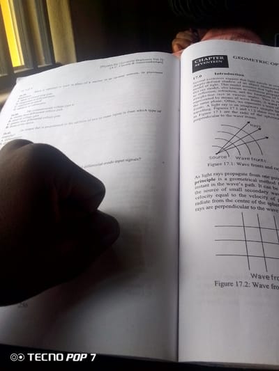 The media file is a close-up, slightly blurry photograph of an open book, likely a textbook, with a hand obscuring a portion of the left page. The book is open to a chapter titled "CHAPTER SEVENTEEN GEOMETRIC OP" and discusses optics, specifically the behavior of light rays and wave fronts. The text on the left page appears to be multiple-choice questions related to electronics, possibly from a physics or engineering course, with question numbers ranging from 16.39 to 16.41. Visible text includes "Physics for University Beginners Vol 2] [A.C. Circuits & Semiconductors]", "16.39.", "16.40.", "16.41.", "CHAPTER SEVENTEEN", "GEOMETRIC OP", "17.0 Introduction", "Source", "Wave fronts", "Figure 17.1: Wave fronts and ray", "256", "Wave fro", "Figure 17.2: Wave fro", and "TECNO POP 7". The setting appears to be indoors, with natural light coming from the left, suggesting it might be daytime. The overall tone is academic and focused on learning. There are no people clearly visible other than the hand holding the book open. The presence of a phone brand logo at the bottom suggests the image was taken with a mobile device.