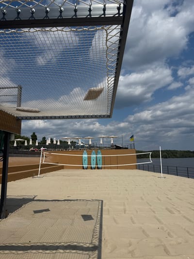 This image captures a recreational area during daytime at Hlibivka, Ukraine. The foreground features a sandy beach, part of which is set up as a volleyball court with a white net and two white poles. Immediately behind the volleyball net, three light blue paddleboards with a textured design are propped vertically against a brown, multi-level terraced structure. On the upper level of this structure, several white sun umbrellas are open, beneath which a few indistinct human figures are visible, appearing to be lounging. A Ukrainian flag, blue and yellow, is flying from a pole on the right side of this elevated terrace. Beyond the terrace, a large body of water extends to a tree-lined horizon. The sky above is partly cloudy with patches of blue. The perspective is from directly beneath an elevated white net structure, possibly a large lounging hammock, supported by a dark metal frame. Two beige cushions are resting on this overhead net, and shadows from the net and cushions are cast onto the sand below in the lower left.