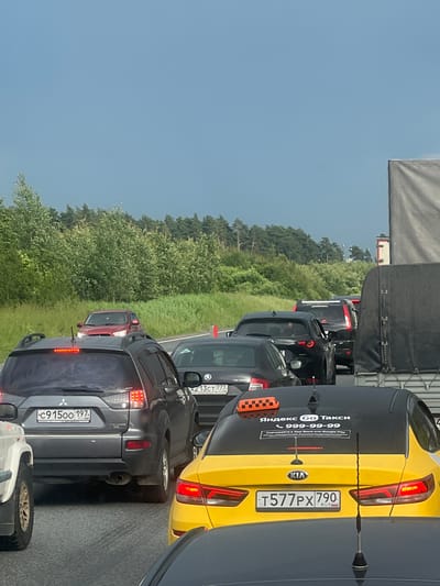 The image depicts a traffic jam on a highway with a dense line of cars. In the foreground, a gray Mitsubishi Outlander is visible with its brake lights illuminated. To its right, a yellow Kia Rio taxi with the Yandex Go logo and phone number "999-99-99" is also stopped in traffic. A black Skoda Octavia with the license plate "A213CT 777" is in front of the Kia. Further down the road, a red SUV is visible, and the highway is bordered by a dense forest and grass. The sky is overcast with a dark cloud formation. The scene is consistent with a traffic jam occurring on a highway near Lukhovitsy, Russia.