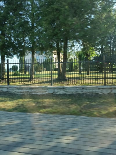 The image depicts a blurred outdoor scene, likely captured from a moving vehicle, showing a fence line, trees, and a building in the background. The fence is made of dark vertical posts and horizontal bars. Beyond the fence, a grassy area with some small trees and bushes is visible. In the middle ground, a light-colored building with multiple windows can be seen, suggesting a residential or institutional structure. The foreground features a gray tiled or paved surface, also appearing blurred due to motion. The overall impression is of a sunny day with shadows cast by the trees on the ground. The specific location in Czartajew, Poland, cannot be determined from the visual information alone. There are no individuals clearly identifiable in the image.