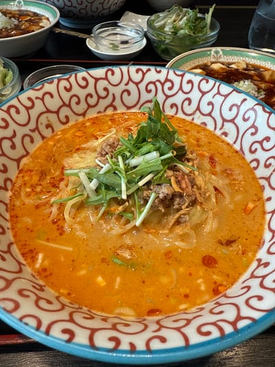 A top-down, slightly angled shot showcases a vibrant bowl of what appears to be a spicy Asian noodle dish, likely ramen or tantanmen, as the central subject. The large ceramic bowl features a distinctive pattern of swirling, interlocking red-orange wave or cloud-like motifs against a white background, with a solid light blue rim.

The bowl is filled with a rich, opaque orange-red broth that looks creamy and slightly oily, with small flecks of a lighter ingredient visible throughout. Submerged within the broth are numerous thin, light-colored noodles.

The dish is generously topped in the center with crumbled, browned ground meat, which appears to be either pork or beef. Mixed with the meat are crispy, golden-brown fried bits, possibly fried garlic or onions, adding texture. A fresh green garnish sits atop the meat, consisting of thin, leafy greens resembling mizuna or arugula, along with shorter, chopped pieces of green onions or scallions.

To the left of the main bowl, several other dishes and containers are visible, suggesting a multi-course meal or a shared dining experience. One plate, partially visible with a green rim, holds a dark, glossy sauced dish, topped with chopped green onions, which could be mapo tofu or a similar braised item. Below it, another bowl with a brown-colored dish is partially seen. Further left, two small, clear glass containers hold shredded green lettuce or a light salad. A small, empty glass bowl with a spoon is also present. The table surface beneath the dishes is dark, possibly black or dark brown.

The lighting is bright and even, highlighting the colors of the food and the details of the patterned bowl, suggesting an indoor setting. No people or visible text are present in the image.