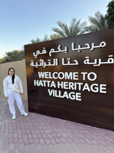 A single young adult female stands on a reddish-brown brick paved walkway next to a large dark brown welcome sign at the Hatta Heritage Village in Hatta, United Arab Emirates. The sign prominently displays "WELCOME TO HATTA HERITAGE VILLAGE" in white raised English lettering, with corresponding Arabic text above it. The female is dressed in a white hooded sweatshirt, white sweatpants, and white athletic shoes, with a light-colored crossbody handbag slung over her right shoulder. She faces the camera with a neutral expression. Behind the sign and the subject, numerous green palm trees are visible under a clear blue sky. To the left of the female, a segment of a light brown building wall is present. The scene is illuminated by natural daylight.