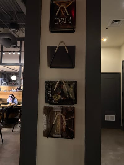A row of four framed items hangs on a white wall with a dark gray ledge above and below. The items include a book titled "DALI", a plain dark gray square, a book titled "THE BEATLES FIFTY YEARS WITH...", and a book titled "UNIVERSITIES THE MOST BEAUTIFUL...". Each item has a U-shaped holder with white wrapped around the middle. Below, a view into a coffee shop is visible with patrons sitting at tables, a counter, and overhead lighting fixtures.