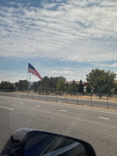 The image depicts an American flag flying on a flagpole on a cloudy day. The flag is positioned to the left of a large building complex that appears to be a corporate office or residential building, surrounded by trees and a chain-link fence. The scene is viewed from a moving vehicle, as indicated by the reflection in the side-view mirror in the lower left corner of the frame. The road in front of the vehicle has white lane markings. The location is in Vacaville, United States of America.
