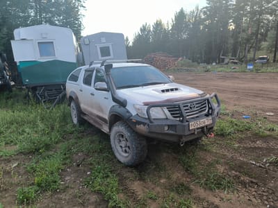 A white Toyota Hilux is parked on a dirt road in a wooded area, possibly near the city of Gremyachiy, Russia. The vehicle is equipped with a snorkel, a bull bar, and aftermarket fender flares. It is covered in mud, indicating it has been driven off-road. To the left of the truck is a white and green trailer with a window, and behind it, another grey trailer with a window. A pile of logs is visible in the background, along with several trees and dense foliage. The ground is a mixture of dirt and grass, with tire tracks visible in the mud. The lighting suggests it is daytime, possibly late afternoon due to the angle of the sun and the hazy quality of the light.