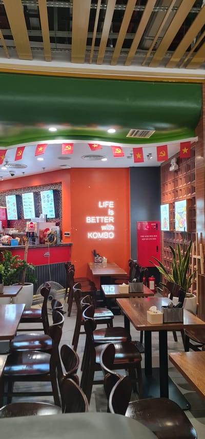 Here is a detailed description of the image:

The image shows the interior of a restaurant in An Cựu Ward, Vietnam. The restaurant has an orange wall with a neon sign that reads "LIFE is BETTER with KOMBO". There is also a fire hose reel box on the wall. The ceiling is decorated with Vietnamese flags.

The restaurant is furnished with wooden tables and chairs. There are plants on the tables, and a fan is visible in the background. There are menus displayed on screens.

The restaurant appears to be empty.