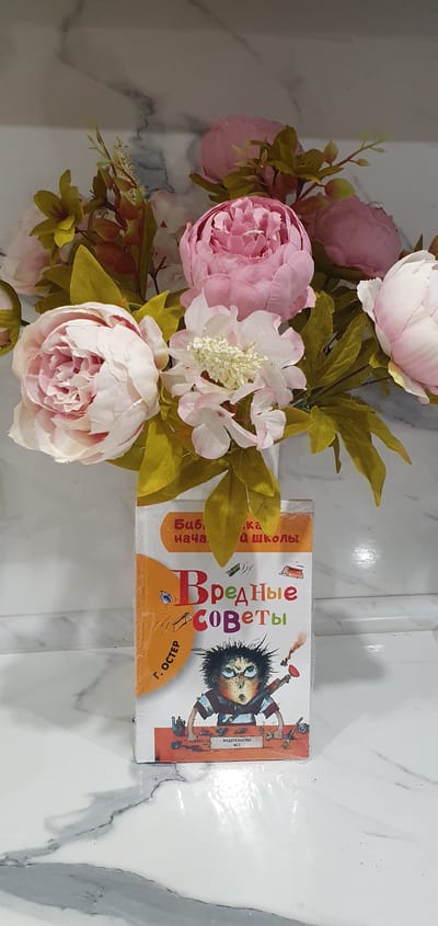 The image shows a bouquet of pink and white flowers next to a book titled "Harmful Advice" in Russian. The flowers are predominantly peonies, with a few other smaller white flowers interspersed. The book has an illustration of a mischievous-looking child on the cover. They are placed on a surface with a white marble pattern. The location is assumed to be Bazarnyye Mataki, Russia, based on the likely language and origin of the book.