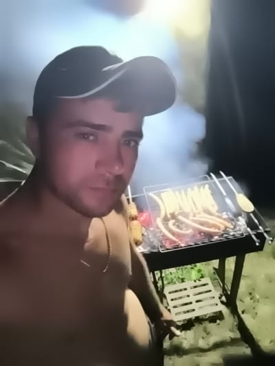 The image depicts a person wearing a dark baseball cap, positioned in the foreground, facing the camera. The individual appears shirtless and is looking directly at the viewer. Behind the person, a grill is set up with numerous sausages cooking over an open flame. Smoke billows from the grill, indicating that the food is actively being cooked. The background is dark, suggesting it is nighttime, with some blurry foliage visible. The scene appears to be outdoors, possibly in a backyard or a campsite. The overall impression is one of a casual outdoor cooking event. The metadata specifies the location as Țipletești, Moldova, though this is not visually discernible from the image itself.