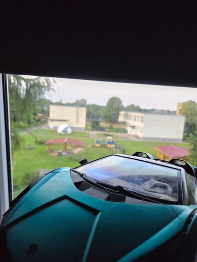 The image shows a close-up view of a teal-colored toy sports car with a clear windshield, positioned on a windowsill. Beyond the windshield, a green grassy yard is visible, with trees and buildings in the background. The yard appears to be a playground or park area, with a red-roofed structure, a white dome-shaped tent, and various colorful play structures. Two multi-story buildings, possibly schools or apartments, are situated further in the distance. The sky is overcast. The toy car's windshield reflects a colorful, possibly iridescent, light. The overall scene suggests an outdoor setting, possibly a schoolyard or community area in Širvintos, Lithuania.