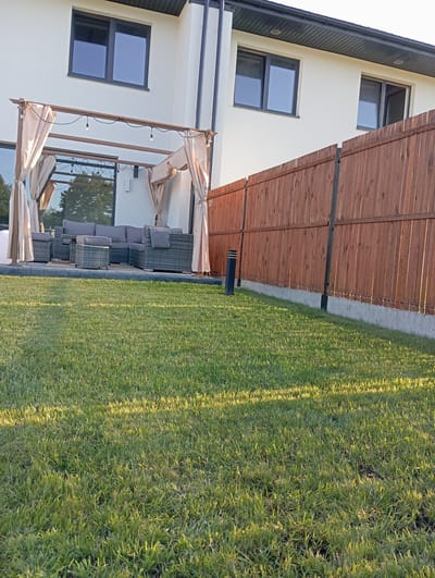 The image shows a backyard scene in Dybów Kolonia, Poland. In the foreground, a well-maintained green lawn is illuminated by sunlight, casting long shadows. To the left, a patio area is visible with a modern outdoor furniture set, including a sectional sofa, armchairs, and a coffee table, all made of dark wicker. A canopy structure with beige drapes covers part of the patio, and string lights are suspended from its frame. Behind the patio, the side of a two-story house with white walls and dark-framed windows is visible. To the right, a tall wooden fence, with dark metal posts, runs along the property line. A dark, modern-looking outdoor light fixture stands in front of the fence. The overall scene depicts a clean and modern residential outdoor space.