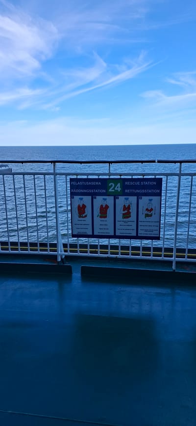 This media file shows a rescue station sign on a ferry. The sign displays instructions for donning a life jacket in four steps, illustrated with diagrams of a person. The text on the sign is in multiple languages, including Finnish, Swedish, and English, with headings such as "RESCUE STATION" and "PELASTUSASEMA". The sign is mounted on a white railing, behind which the sea is visible. The sea appears calm with ripples. The sky above is a bright blue with wispy white clouds. The overall lighting suggests it is daytime, likely a clear and pleasant day. The ferry deck, visible on the right side of the image, is a dark blue color. The scene appears to be a standard safety procedure display on a vessel, offering no specific emotional cues beyond the inherent seriousness of safety instructions.