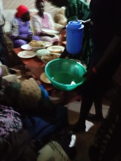 A social gathering shows people seated near a table laden with plates and food. A blue water jug sits prominently on the table. A large green bowl is in the foreground. People are standing. The lighting is dim, making details indistinct. Clothes and bags are on the floor.