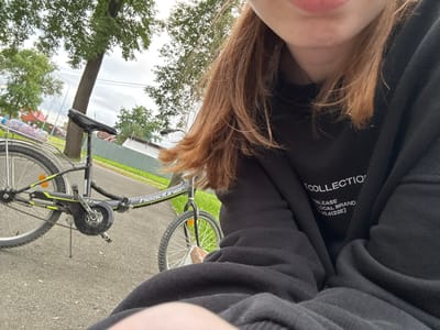 A close-up, low-angle, selfie-style shot shows a person's head and shoulder, partially obscured by their long, reddish-brown hair and a black hooded sweatshirt. The person appears to be outdoors, with a paved surface in the foreground and green grass and trees in the background. A bicycle is visible, lying on its side, with its wheels and drivetrain in focus. The bike is black with green and yellow accents. The background suggests a slightly overcast day, with bright skies and lush green foliage. A thin wooden post with a shoe dangling from it is also visible. Text is visible on the inside of the sweatshirt, reading "COLLECTION," "RELEASE," "LOCAL BRAND," and "(60.6122E)". The overall mood is casual and relaxed, suggesting a moment of rest or a break during an outdoor activity. The text and the bike suggest this is a candid shot taken by a person out and about in Nevyansk, Russia.