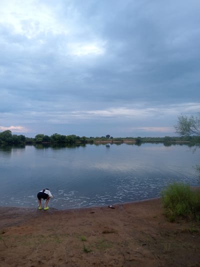A young person, wearing a white t-shirt and black shorts, is seen from behind in a crouching position at the edge of a wide river. They are wearing bright yellow shoes. The river has a calm surface with gentle ripples, and on the far bank, a dense line of green trees is visible, reflected in the water. The sky above is overcast with muted blue and grey clouds. The foreground consists of a sandy shore with scattered debris, including a pink object near the water's edge. The scene appears to be in a natural setting, likely a park or undeveloped area near the river. The location is identified as Vasil'evka, Russia.