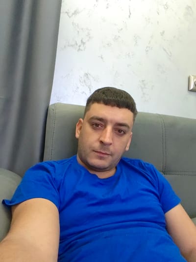 A male individual is depicted in a close-up photograph, seated indoors in Opaka, Bulgaria. The adult male subject has short dark hair with a straight fringe and closely shaved sides, and a neutral facial expression with his gaze directed towards the camera. He is wearing a royal blue short-sleeved t-shirt. The subject is positioned against the backrest of a gray upholstered sofa or armchair. To the left, a dark gray curtain is partially visible. The wall behind the subject is light-colored with a mottled gray, textured finish. A light-colored electrical outlet or switch plate is affixed to the wall in the upper right quadrant of the image.