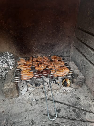 In the city of Ayvacık, Turkey, a barbecue grill is filled with pieces of chicken. The grill is positioned over hot coals within a rustic fireplace. To the left of the grill, a lump of food wrapped in aluminum foil sits on a brick. The fireplace walls are made of rough, dark stone, and the ground is covered in ash and small pieces of wood. An onion, partially charred, rests on the coals to the right of the grill. The scene captures the process of cooking outdoors over an open fire.