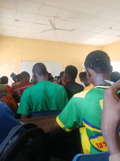 The scene depicts an indoor classroom or lecture hall in Ugi Majowel, Nigeria, viewed from behind the seated attendees. The room features light yellow walls and a white tiled ceiling with a three-bladed white ceiling fan centrally mounted. A whiteboard with faint written content is visible on the far wall. Natural light illuminates the room from a window on the right side.
Approximately eight individuals, primarily appearing as young adults, are visible in the foreground and midground, seated at desks and facing forward. In the immediate right foreground, the back of an individual wearing a green and yellow striped short-sleeved shirt with a red graphic on the rear is seen, with their right hand holding a writing instrument. To their left, another individual wears a plain green short-sleeved shirt. Other attendees are dressed in various casual attire, including one person in a patterned brown and red shirt. A black or dark blue backpack rests on a desk in the lower-left foreground. Several individuals in the mid-ground are holding up mobile phones, directed towards the front of the room, while another individual on the left holds a blue object, possibly a book or tablet. The individuals are positioned as if observing a presentation or lecture.
