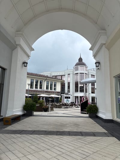 This is a vertical, eye-level, full shot of an arched walkway leading into a plaza. The archway is white and ornate, with decorative molding on the underside and sides. Beyond the archway, a plaza is visible with paved ground, outdoor seating areas with tables and chairs, and umbrellas. In the background, there are several buildings. The building directly ahead has a prominent dome with a spire on top. To the left, a longer building features a sign that reads "SHORE HOUSE VILLAGE". The sky is overcast. A wooden bench is situated to the left of the archway. The environment appears to be a commercial or entertainment district, possibly part of Millennium Park in Russia, characterized by its modern architecture and outdoor amenities. No people are visible in the image.