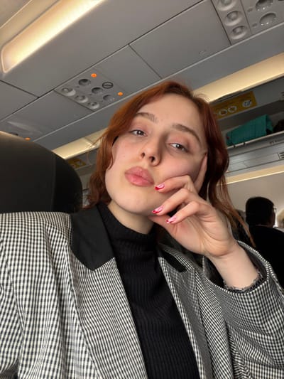 The image captures a female individual inside a commercial aircraft cabin. The scene is contextually placed in Tuzla, Turkey.

**People:**
A young adult female is centered in the frame, directly facing the camera in a self-portrait. She possesses light skin, reddish-brown hair, and dark eyes. Her lips are slightly puckered in what appears to be a "duck face" expression. Her left hand is raised, with fingers gently touching her chin and cheek. Her nails are painted light pink with distinct red wavy or heart-like designs. A silver-colored bracelet is visible on her right wrist. In the background on the far right, the top of another passenger's head, with dark hair, and shoulder are partially visible, facing away from the camera.

**Subjects/Objects:**
The primary subject is the female individual. She is wearing a black, ribbed turtleneck top under a black and white small-checkered blazer. The blazer features solid black lapels.
The immediate environment is a commercial airplane interior. Above the subject's head, the gray overhead passenger service unit is visible, equipped with individual reading lights, air vents, and call buttons; some indicator lights appear orange. Overhead bin doors are present, one with a yellow safety sticker and another containing a teal-colored bag. An illuminated strip light runs along the cabin ceiling. A black airplane seat headrest is partially visible on the left.

**Actions/Interactions:**
The female individual is engaging in a self-portrait, holding her left hand to her face in a casual pose. No direct interaction with other individuals is depicted.

**Environment:**
The environment is an airplane cabin, characterized by standard overhead panels, lighting, and seating arrangements of a commercial airliner. The interior appears clean and well-lit by both cabin lighting and an ambient glow from an unseen source (possibly a window or additional cabin lights).