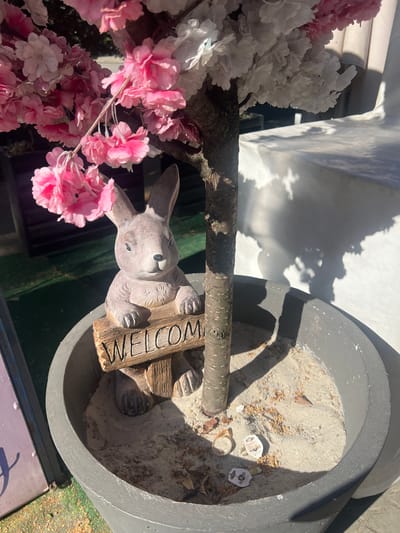 In Berehove, Ukraine, a close-up shot captures a decorative ceramic bunny holding a wooden sign that reads "WELCOME." The bunny and sign are situated in a large gray planter filled with sand and the base of a faux cherry blossom tree, adorned with pink and white flowers. The planter is placed outdoors, with dappled sunlight casting shadows from the tree onto a white surface in the background. Scattered within the sand are a few dried leaves and what appear to be discarded price tags.