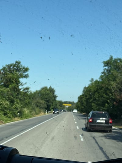 The image depicts a road scene from the perspective of a vehicle's interior, looking through the windshield. The windshield has numerous smudges and specks, obscuring the view slightly. The road is asphalt and appears to be a two-lane highway with white lane markings. On the right side of the road, a black car is driving in the same direction. Trees line both sides of the road, with lush green foliage. The sky is a clear blue with some scattered lighter patches. In the distance, there appears to be a yellow sign or structure. The scene suggests a sunny day, likely in a rural or semi-rural area, possibly on the outskirts of Osenets, Bulgaria, given the context. There are no people visible, and the focus is on the road and surrounding environment.