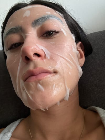 The media file is a close-up, vertically oriented photograph of a person's face adorned with a sheet mask. The subject appears to be a woman with dark hair, her face is angled slightly upwards. She is lying down, with her head resting on a dark, textured surface, possibly a couch or pillow. A light-colored garment, possibly a sweater or robe, is visible near her shoulder and neck. A thin, gold chain necklace is also visible around her neck.
The sheet mask is white and translucent, with visible moisture or serum. It covers most of her face, with openings for the eyes and mouth. Her eyes are open and looking upwards, and her lips are slightly parted. The lighting is soft, suggesting an indoor setting. There are no visible text elements in the image. The overall mood appears relaxed and focused on self-care. No specific time of day or weather conditions are discernible. The provided location context of Imatra, Finland, does not offer any visual cues within the image itself.