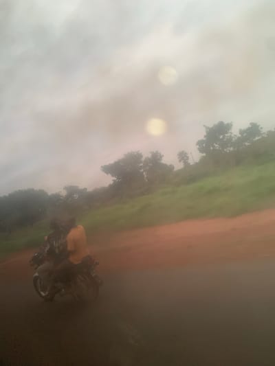 The image depicts two individuals on a motorcycle traveling on a paved road. The person in the back is wearing a yellow shirt, while the person in the front is wearing a dark patterned shirt. They are positioned on the left side of the road. The environment surrounding them features a grassy hill on the right, with several trees scattered across it and a dirt shoulder separating the road from the hill. The sky is overcast with visible light flares, suggesting a hazy or foggy atmosphere. The location is identified as Nev, Nigeria.