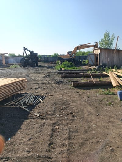 The image depicts an outdoor scene at a construction or lumber yard, likely in Strelka, Russia, based on the context. The foreground is dominated by a rough dirt surface with scattered debris. To the left, a large stack of finished lumber is prominently displayed, with smaller pieces of wood scattered in front of it. In the middle ground, a dark-colored logging truck with a crane attachment is parked. Further to the right, a yellow Hyundai excavator is positioned near a rustic wooden shed with an open door. Several large logs are arranged on the ground between the excavator and the shed, and more lumber is visible leaning against the shed. The background features a wooden fence and vegetation, with a clear blue sky overhead. No people are visible in the frame.