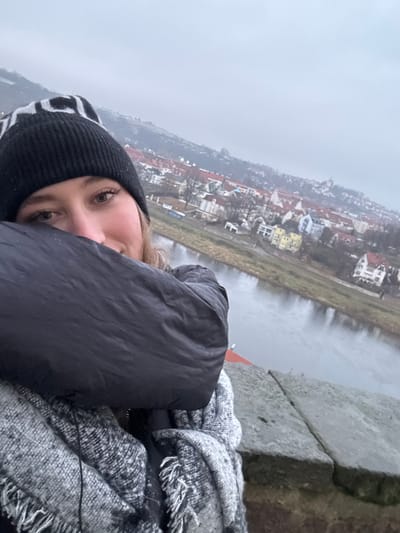 An image, captured in portrait orientation and rotated 90 degrees counter-clockwise, features a young woman in the foreground against a scenic backdrop of a European town and river.
The woman is positioned on the left side of the frame (when viewed correctly), with her face partially obscured by her dark puffer jacket, revealing one eye and a portion of her nose and cheek. She is wearing a black knitted beanie with a white pattern and a textured grey and white scarf with fringe, suggesting cold weather. Her visible eye looks directly at the viewer, indicating a selfie or a close-up photograph.
The background showcases a river flowing through a valley, bordered by a town with numerous buildings featuring diverse roof colors (red, white, yellow) and light-toned facades. Beyond the town, the landscape is hazy and atmospheric, hinting at distant hills or urban areas under an overcast, grey sky. A robust stone wall or parapet is visible on the right side of the image, suggesting the vantage point is from a raised structure overlooking the river and town, likely in Meissen, Germany. The overall atmosphere is cool and cloudy, consistent with the heavy clothing worn by the subject. No discernible text is visible in the image.
