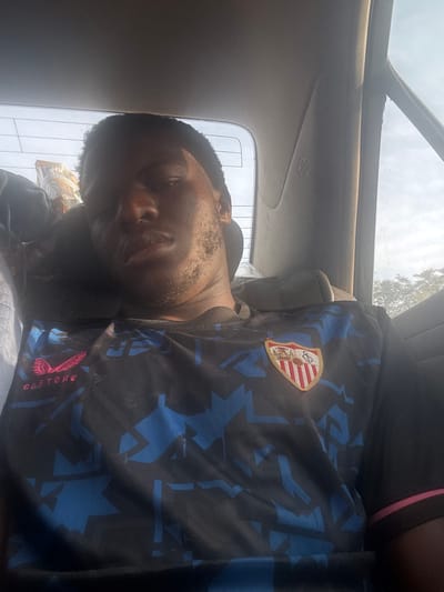 A man is reclined in a car seat, appearing drowsy or asleep. He is wearing a dark sports jersey with a prominent crest or logo on the chest and blue geometric patterns. The word "CASTORE" is visible on the shirt. The car's interior includes a sunroof or window above. The lighting is somewhat harsh, with some areas overexposed.