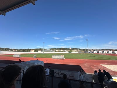 The image depicts a daytime scene at a soccer stadium in Valpaços, Portugal. A red running track surrounds a green soccer field, which is being watered by sprinklers. Several covered player benches are visible near the track. In the background are houses and trees on a slight hill, with a religious building visible on the peak.
In the foreground, several people are partially visible, suggesting the image was taken from the stands. A banner with the words "Otica flavea," "aria Juvenal II", and phone numbers is attached to the stadium fence.