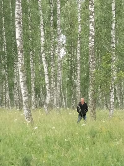 The image shows a man standing in a grassy clearing surrounded by a dense forest of birch trees. The man is wearing a black jacket and blue jeans. He is facing the camera with his hands in his pockets. The grass in the clearing is tall and green, with some small white flowers scattered throughout. The birch trees are tall and slender, with characteristic white bark and green leaves. The forest appears to be in a rural area. The location is the city of Idrisovo, Russia.