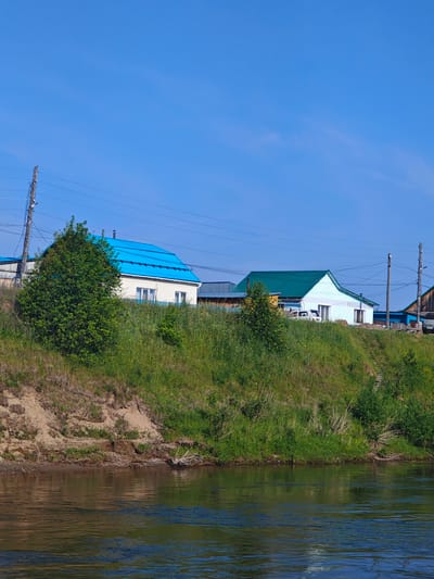 The image depicts a sunny day scene in Galanino, Russia. In the foreground, a calm river flows, reflecting the sky and surrounding greenery. The riverbank is a steep, grassy slope with exposed soil in some areas, and sparse vegetation.
Further up the slope, a small village is visible. Several houses with distinctively colored roofs, one bright blue and another green, are nestled among trees and tall grass. Power lines stretch across the sky, supported by wooden poles. The houses appear to be residential, with windows and doors visible. A white car is parked in front of the house with the green roof. The overall environment is rural and serene, with no people present in the frame. The sky is a clear, bright blue.