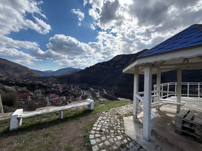 The image captures a scenic overlook in the city of Bov, Bulgaria. A white wooden gazebo with a blue roof is positioned on the right side of the frame. Inside the gazebo, a bench is visible along with a makeshift wooden pallet table.
Outside the gazebo, to the left, stands a weathered white bench. A cobblestone path connects the bench to the gazebo.
In the background, a small town nestled in a valley is visible. The town consists of buildings with red roofs. Behind the town, forested hills and mountains rise into the sky, which is partly covered with clouds. The sky is a mix of blue and grey, with the sun breaking through the clouds. A river and a road can be seen winding through the landscape.