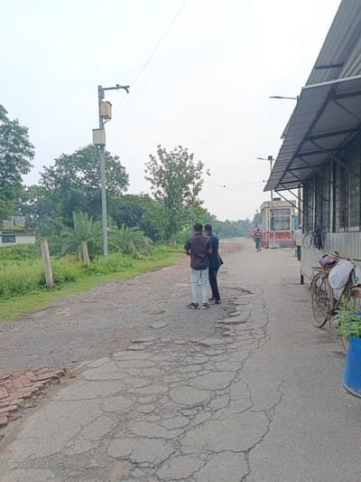 The image depicts a street scene in Benapole, Bangladesh. The road surface is visibly deteriorated with extensive cracking and potholes. On the left side of the road, there is a grassy area with some concrete posts and lush green vegetation. A concrete building is partially visible in the background. In the mid-ground, two men are standing on the road, facing away from the camera. Further down the road, other individuals are walking, including one person in a striped garment.

On the right side of the road, a corrugated metal structure extends into the frame, likely a shop or warehouse with windows and exposed metal framing. A rusted bicycle with a white bag attached to the handlebars is parked next to the structure. A blue plastic bin is also present. Overhead, electrical wires are strung across the scene, and a utility pole with what appears to be electrical boxes is visible on the left. The sky is overcast.