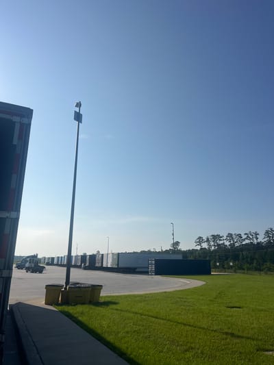 A low-angle, wide shot captures an outdoor industrial setting under a clear blue sky. In the foreground, the side of a dark-colored shipping trailer is visible on the left, with red and white reflective stripes. Adjacent to this trailer, a gray concrete walkway leads to three yellow trash receptacles grouped together. Beyond the walkway and trash bins, a vast expanse of green grass stretches out, bordered by a curved asphalt lot.
On the asphalt lot, a line of identical shipping containers, painted various colors including white, gray, and dark blue, are parked in a row. Further down the lot, a dark gray truck cab is visible, appearing to be maneuvering. A tall, slender light pole stands prominently in the middle ground, with a square fixture at its top. In the distance, a dense line of pine trees forms a treeline against the horizon, with a few buildings and other light poles interspersed. The overall scene suggests a logistics or distribution center in a rural or suburban area, likely during daytime. The location is identified as Pooler, United States of America.