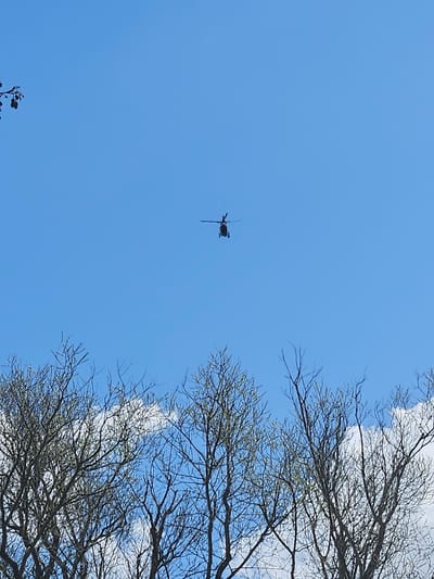 The image shows a helicopter flying in a clear blue sky with some wispy clouds. Bare tree branches are visible on the right side of the frame. The helicopter appears to be in flight over Gavisse, France. The helicopter is distant and silhouetted against the sky.