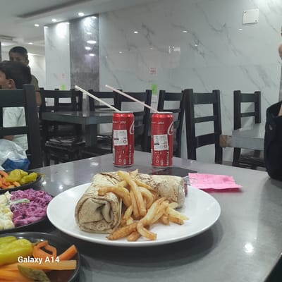 Here is a description of the image:

The image depicts a table setting in what appears to be a restaurant, possibly located in Nuseirat, Palestinian Territories. On the table, there's a white plate containing a wrap (possibly shawarma) accompanied by fries. Two cans of Coca-Cola with straws sticking out are placed near the plate. Additionally, there are side dishes in black bowls, seemingly containing pickled vegetables or salads. In the background, there are black chairs and glimpses of people, suggesting the restaurant is open and serving customers. The setting looks like a casual dining environment. A pink sticky note is on the table. "Galaxy A14" is written on the picture.
