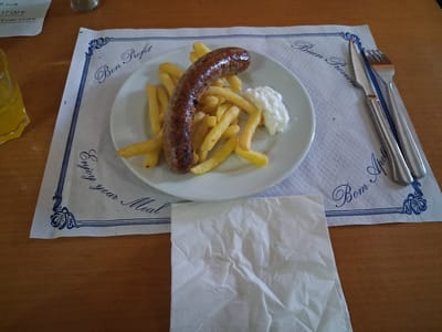 The image shows a plate of food on a table in what appears to be an outdoor cafe setting in Mas Aliu, Spain. The main subject is a meal consisting of a sausage and french fries served on a white plate. Next to the food is a dollop of white sauce, likely aioli or mayonnaise, a common accompaniment to french fries in Spain. The plate rests on a decorative paper placemat with blue lettering. Text visible on the placemat includes "Bon Profit," "Enjoy your Meal," and "Buen Provecho," all phrases wishing the diner a good meal. There is also a white paper napkin folded to the right of the plate. The table surface is made of wood, and a glass with yellow liquid, possibly orange juice or a similar beverage, is partially visible on the left side of the image. The lighting suggests it is daytime. The scene conveys a relaxed, casual dining experience. Visible text on a menu in the upper left corner of the image includes prices, "17.00 €" and "12.00€," and items like "ll-i-oli" and "Y café".