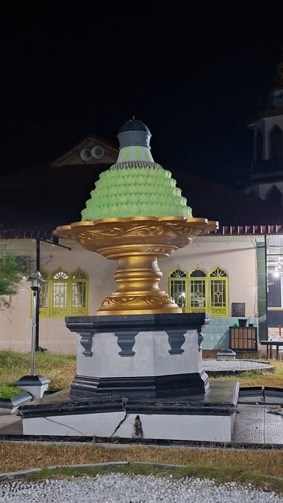 The image depicts an outdoor night scene at a location in Lhokdagang, Indonesia. The central subject is a decorative fountain structure, adorned with a prominent gold pedestal topped with a tiered, light green structure resembling layered leaves or petals, capped with a dark finial. The base of the fountain is a substantial, multi-tiered pedestal with grey and black detailing, showing visible cracks and damage.

In the background, a building with a red tiled roof is visible, featuring arched windows with yellow frames and decorative grilles. Two loudspeakers are mounted on the roofline of this building. To the right, a minaret of a mosque or similar religious building can be seen, with intricate detailing and illuminated sections.

The ground around the fountain is covered with patches of dry grass and gravel. The scene is illuminated by artificial lighting, highlighting the fountain and the architecture in the background. No people are present in the image. The overall atmosphere is serene and architectural, emphasizing the monumental fountain and its surroundings in a nighttime setting.