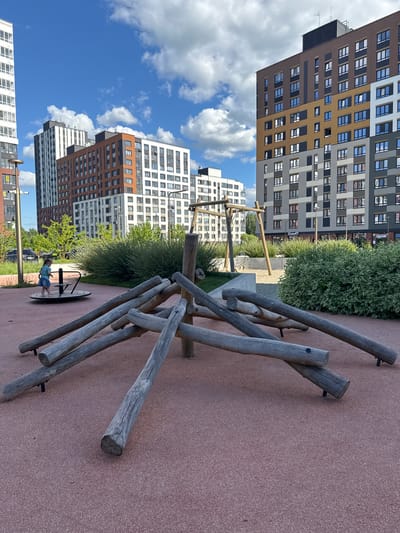 This media file depicts an outdoor scene in a residential area in Patrusheva, Russia. The focal point is a playground with a wooden climbing structure made of rough-hewn logs, angled and bolted together. In the foreground, the ground is a reddish-brown, textured surface, likely a rubberized safety material common in playgrounds.
In the mid-ground, a child is visible on a merry-go-round, appearing to be enjoying themselves. Behind the playground equipment, lush green bushes and trees provide a natural backdrop. Further in the background, modern, multi-story apartment buildings dominate the skyline. These buildings feature a distinct architectural style with alternating colored facades – brown, white, and grey – and numerous windows.
The sky is a vibrant blue with scattered white, fluffy clouds, suggesting a bright, pleasant day. The sun is likely high in the sky, indicating it is midday. The overall mood of the image is peaceful and suggests a family-friendly environment. There is no visible text in the image, aside from a small sign on one of the buildings that reads "СТАРТ." The image is oriented vertically, as if taken with a phone held in portrait mode.