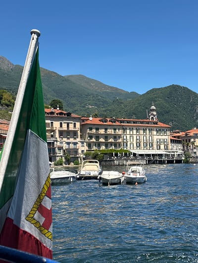The image captures a scenic view from a boat on a body of water, likely a lake, with the Italian flag in the foreground. In the background, a town is visible, characterized by buildings with red-tiled roofs and an array of balconies. These structures are nestled against a backdrop of lush green, tree-covered mountains under a clear blue sky. Several small boats are moored near the shore in front of the town. The water is choppy, reflecting the bright sunlight. The location appears to be in Loveno, Italy, a town known for its picturesque setting on the Italian lakes.