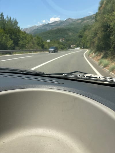 This media file is a first-person perspective video taken from inside a vehicle, likely a car, driving along a paved road. The road has a white dividing line and is flanked by a guardrail on the left. To the left of the road, a steep, tree-covered mountain slopes upwards, with a clear blue sky visible above. In the distance, on the road, another dark-colored vehicle can be seen. The right side of the frame is dominated by the interior of the car, showing a textured dashboard. The image is oriented vertically, suggesting the car is moving forward. The time of day appears to be daytime, with bright sunlight casting shadows. The weather seems clear and sunny. The scenery suggests a mountainous region, consistent with the provided location context of Golubovići, Montenegro. There is no visible text in the media file.
