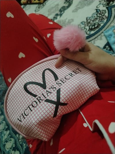 A hand holds a pink, fluffy pom-pom above a Victoria's Secret eye mask. The mask is pink with a grid pattern and the brand logo in black. The person is wearing red pajamas with white hearts. The scene is set against a bed with a patterned blue and white blanket.
