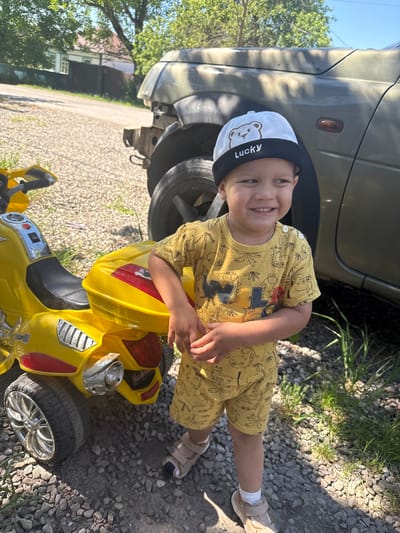 A young boy, likely under five years old, stands outdoors in front of a damaged vehicle and a yellow toy motorcycle. He is wearing a yellow t-shirt with a dinosaur print and matching yellow shorts. On his head, he has a white and black baseball cap with a bear image and the word "Lucky" printed on it. The boy is smiling and looking to his right. The environment appears to be a rural area or the outskirts of a town, with gravel on the ground and some vegetation. In the background, there's a house and trees, suggesting a residential setting. The car appears to be an older model, with its front bumper and some other parts missing or damaged. The yellow toy motorcycle is detailed, resembling a larger motorcycle with a seat, handlebars, and wheels. The lighting indicates it is daytime, likely sunny, based on the shadows. The overall scene is casual and appears to be a snapshot of a child's playtime outdoors. The location is identified as Vinsady, Russia.
